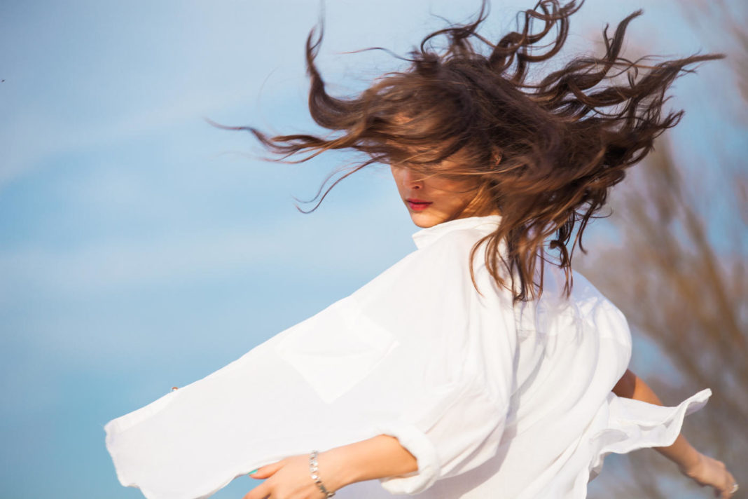 jeune femme dans une chemise blanche qui se tourne avec les cheveux en mouvement, en extérieur, le ciel bleu en fond
